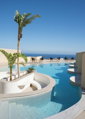 Zona de piscina de disseny corbat, amb gandules i palmeres, amb vista a un cel blau clar i l'oceà.