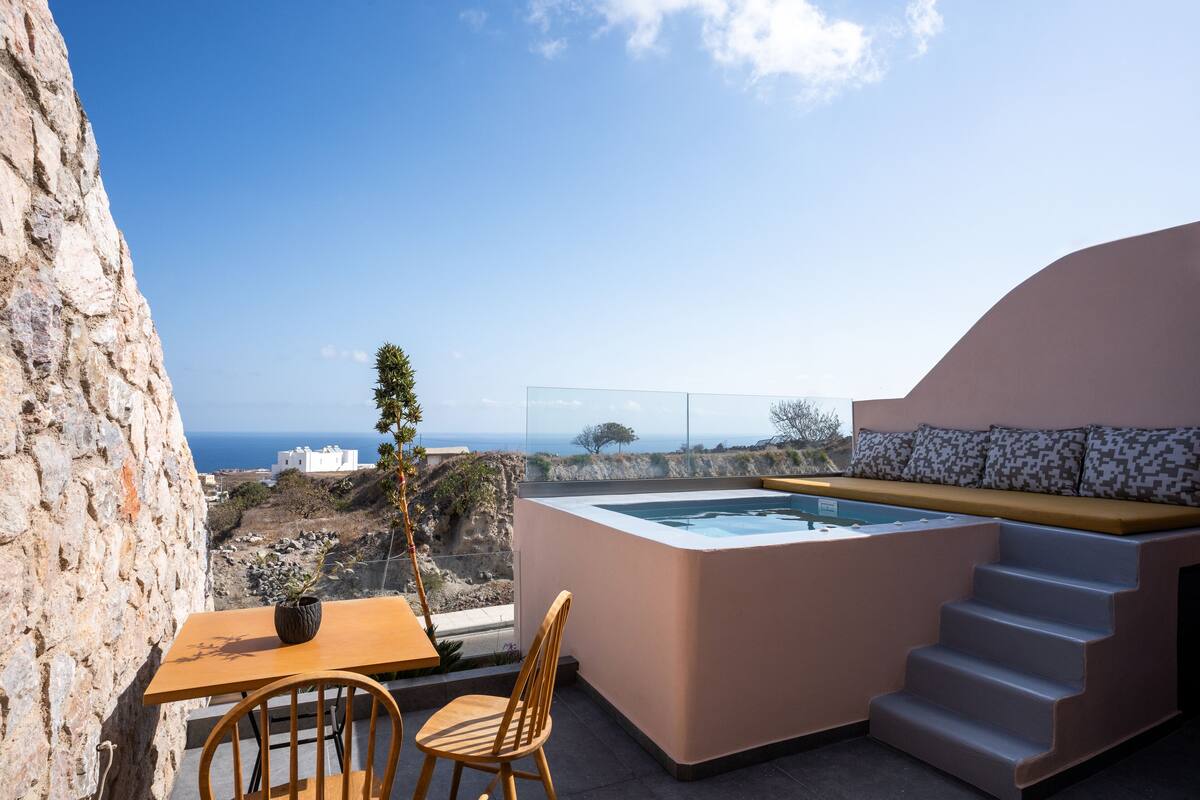 A terrace with a hot tub, a wooden table, and a chair, overlooking a rocky landscape and the sea.
