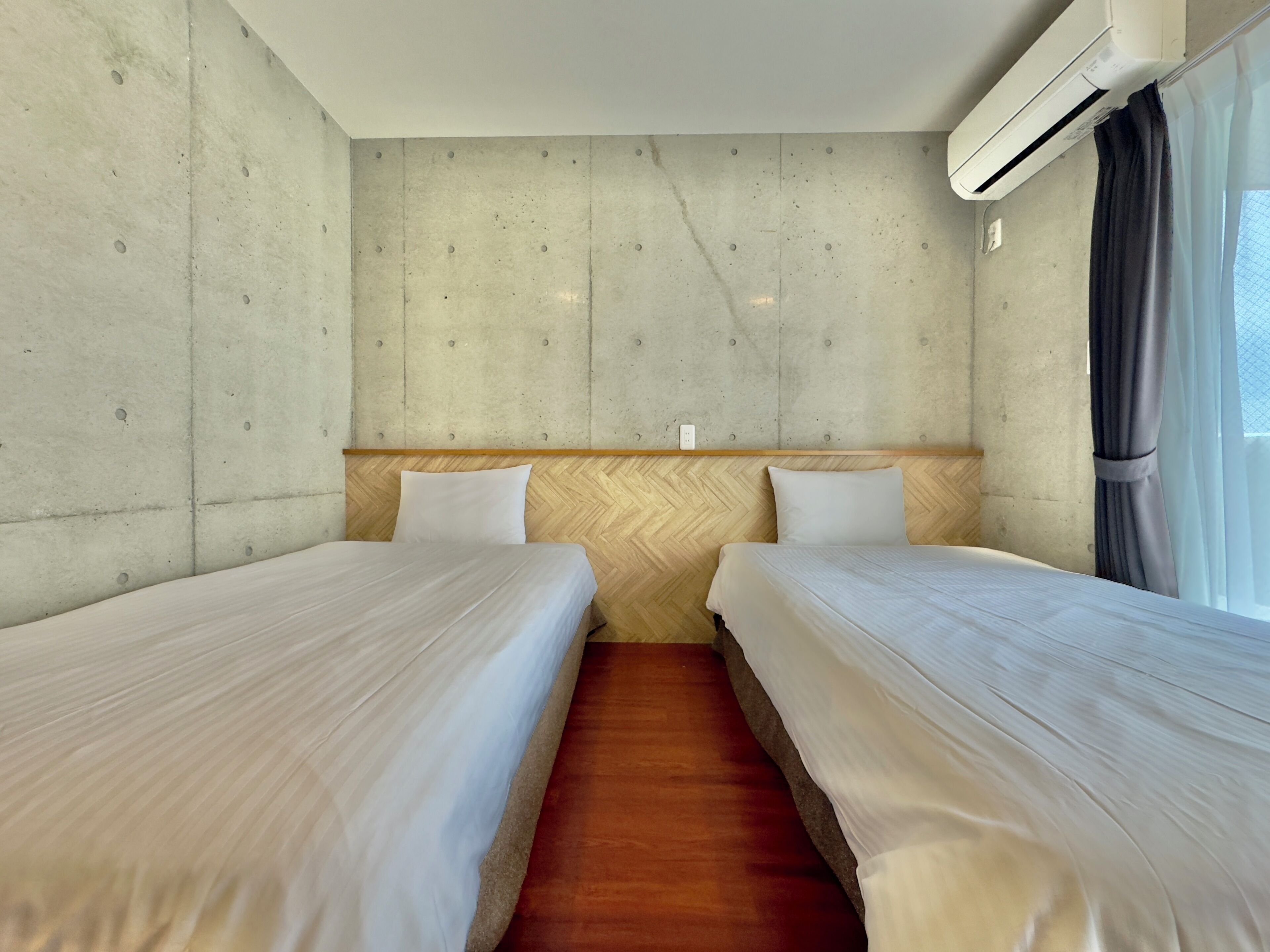 Une chambre d’hôtel avec deux lits simples, une tête de lit en bois, un mur en béton, un appareil de climatisation et une fenêtre avec des rideaux.