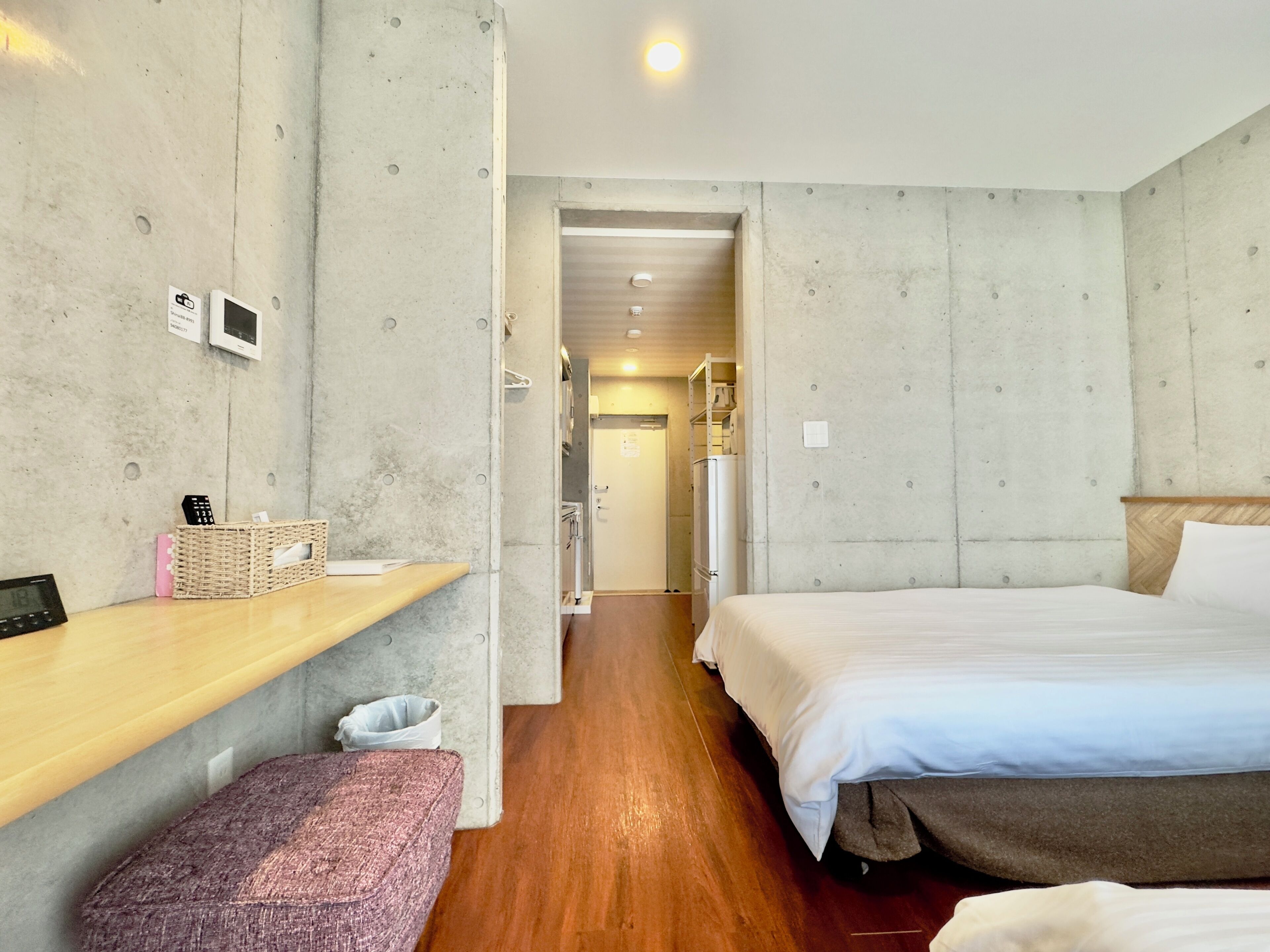 A hotel room with a wooden floor, a bed with white bedding, a bench, a small table with a basket, and a wall-mounted shelf.