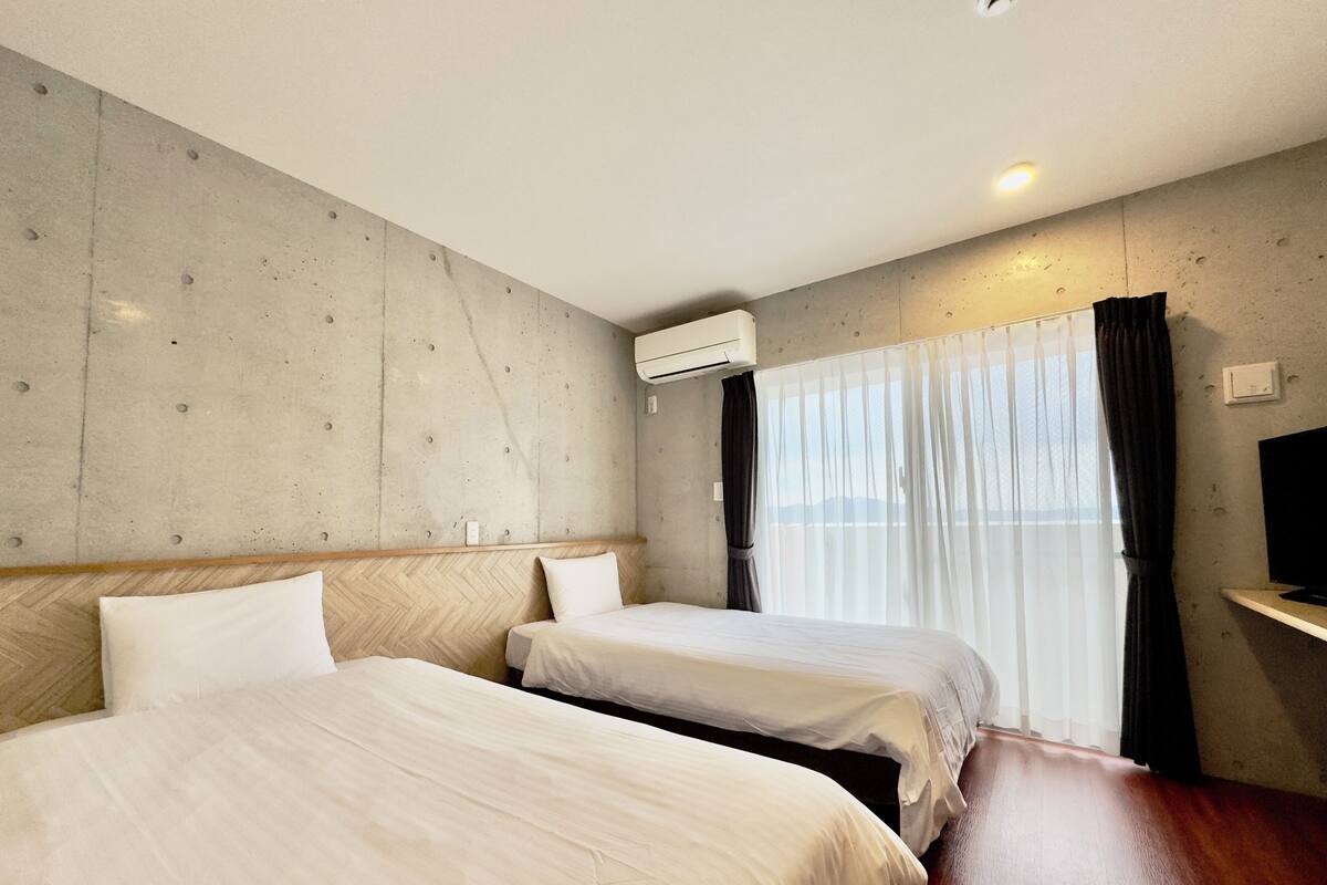 Ein Hotelzimmer mit zwei Betten, einem hölzernen Kopfteil, einem großen Fenster mit durchsichtigen Vorhängen und einem Flachbildfernseher auf einem Regal.