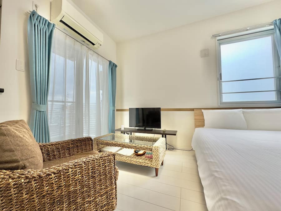A hotel room with a bed, a television on a stand, a wicker chair, and a window with curtains.