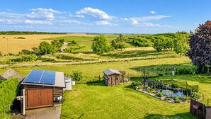 A rural landscape with a house featuring a solar panel, a small shed, and a garden with a pond.