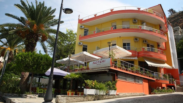 A multi-story building with balconies, a sign reading "NOTERI," and outdoor seating with umbrellas. - Hotel Alpine (Vlorë)