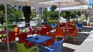 An outdoor seating area with colorful plastic chairs and tables, umbrellas, and a view of the street and buildings in the background. - Hotel Alpine (Vlorë)