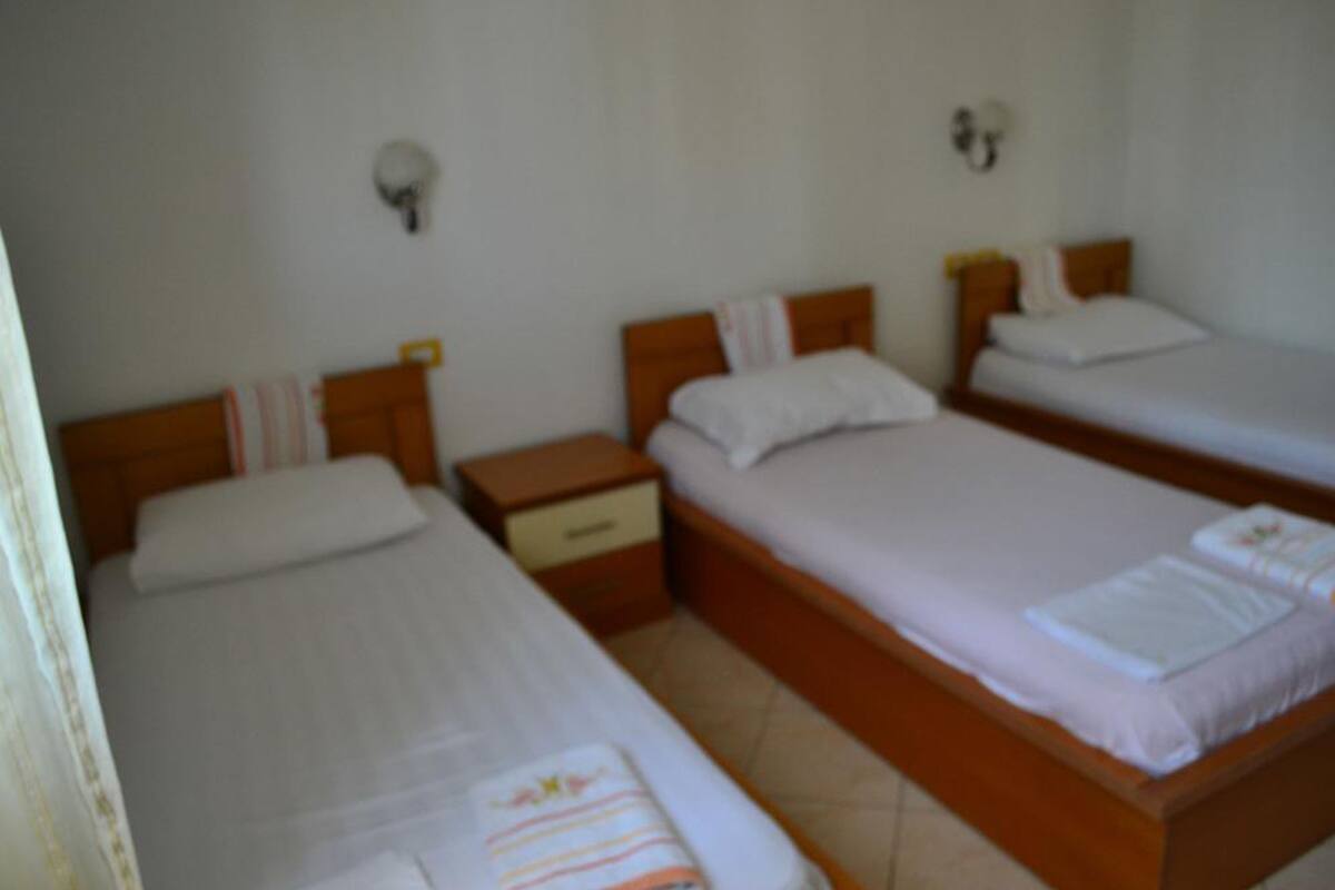A hotel room with three single beds, each with a wooden headboard and white bedding. There are bedside tables with lamps and a small drawer.