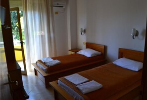 A room with two single beds, each with a wooden headboard and white pillows. There is a small table and a chair to the left, and a window with curtains.