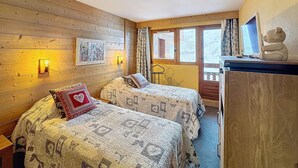 Quarto aconchegante com paredes de madeira, duas camas com colchas estampadas, uma escrivaninha com computador e uma janela com cortinas.
