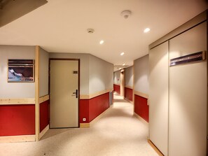Un couloir aux murs rouge et beige, une porte sur laquelle est apposée une pancarte, et un passage qui mène à une autre pièce.