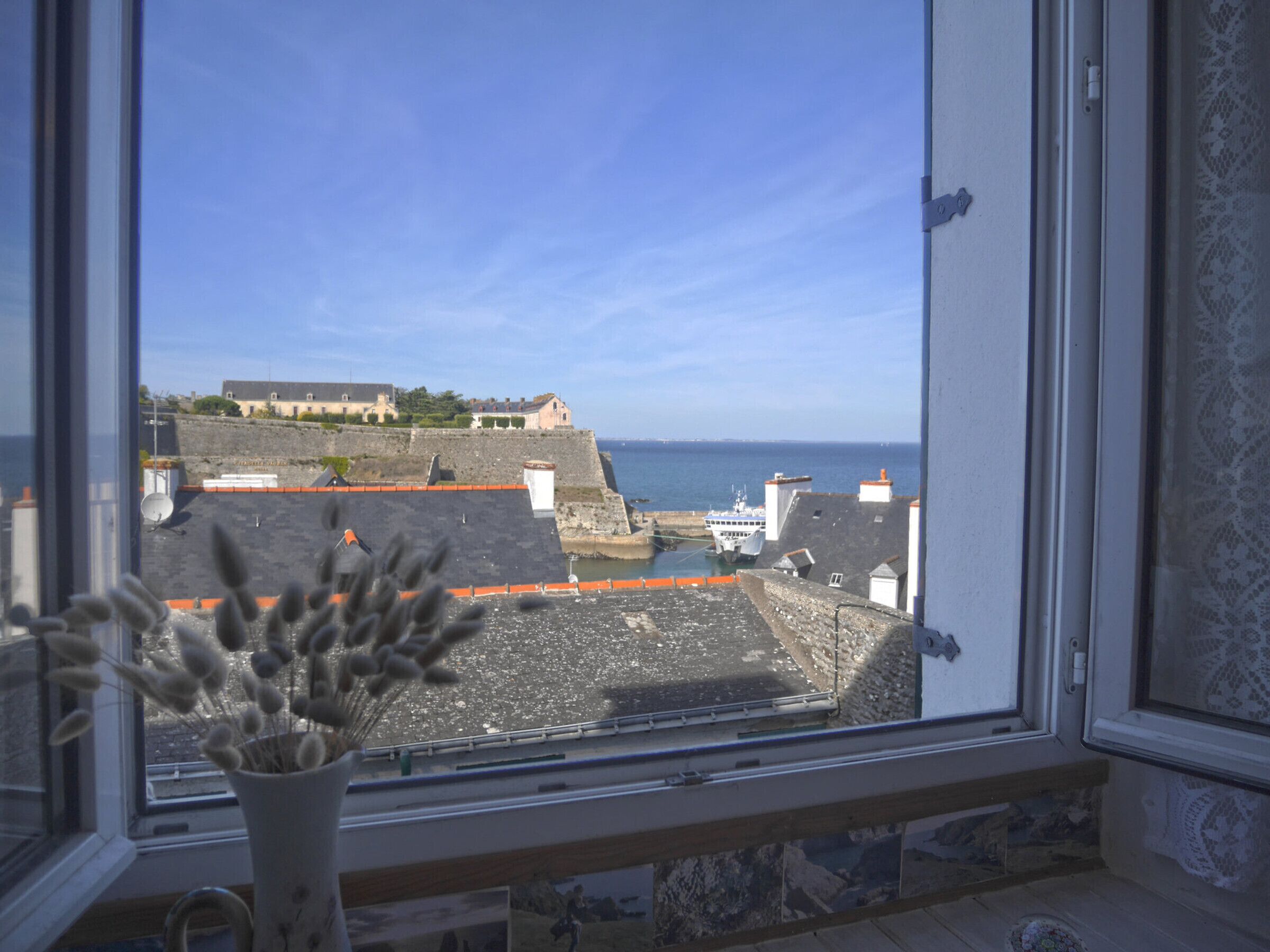 Blick durch ein Fenster auf eine Küstenstadt mit Gebäuden, einer Hafenmauer und einem großen Gebäude auf einem Hügel.