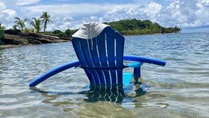 Une chaise bleue, en partie immergée dans une eau claire, recouverte d’un tissu blanc, se trouve au milieu d’un paysage tropical, avec des palmiers et une île visible au loin.