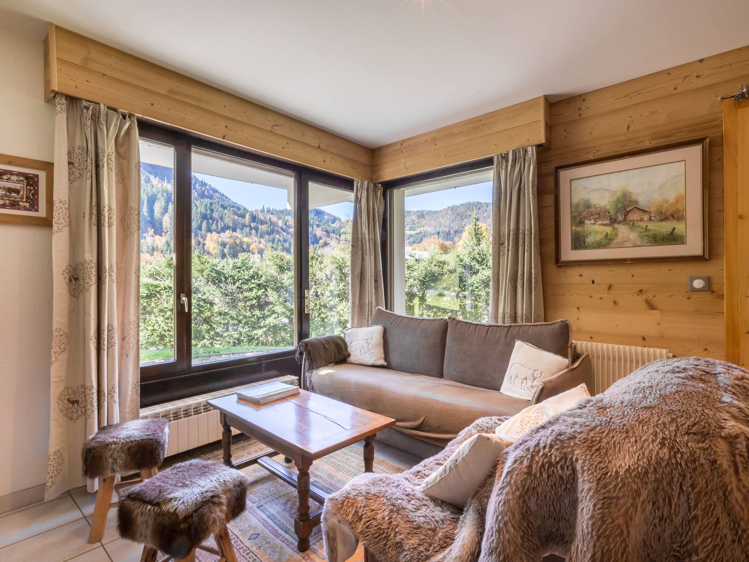 A cozy room with a wooden interior, a sofa, a table, and a view of a forested landscape through large windows.