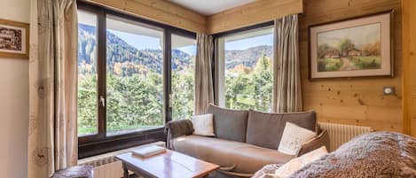 A cozy room with a wooden interior, a sofa, a table, and a view of a forested landscape through large windows.