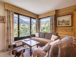 Una habitación acogedora con detalles de madera, un sofá, una mesa y vistas a un paisaje boscoso a través de amplias ventanas.
