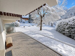 A snow-covered landscape with a building entrance, a tree, and a snow-covered bush.