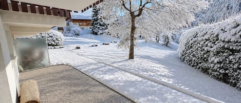 A snow-covered landscape with a building entrance, a tree, and a snow-covered bush.