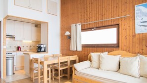 A compact room with a wooden floor, a wooden sofa with cushions, a wooden dining table with chairs, a small wooden table, a kitchenette with a coffee machine, and a window with a curtain. - Tignes Apartment, 2 rooms, 500m from slopes (Tignes)