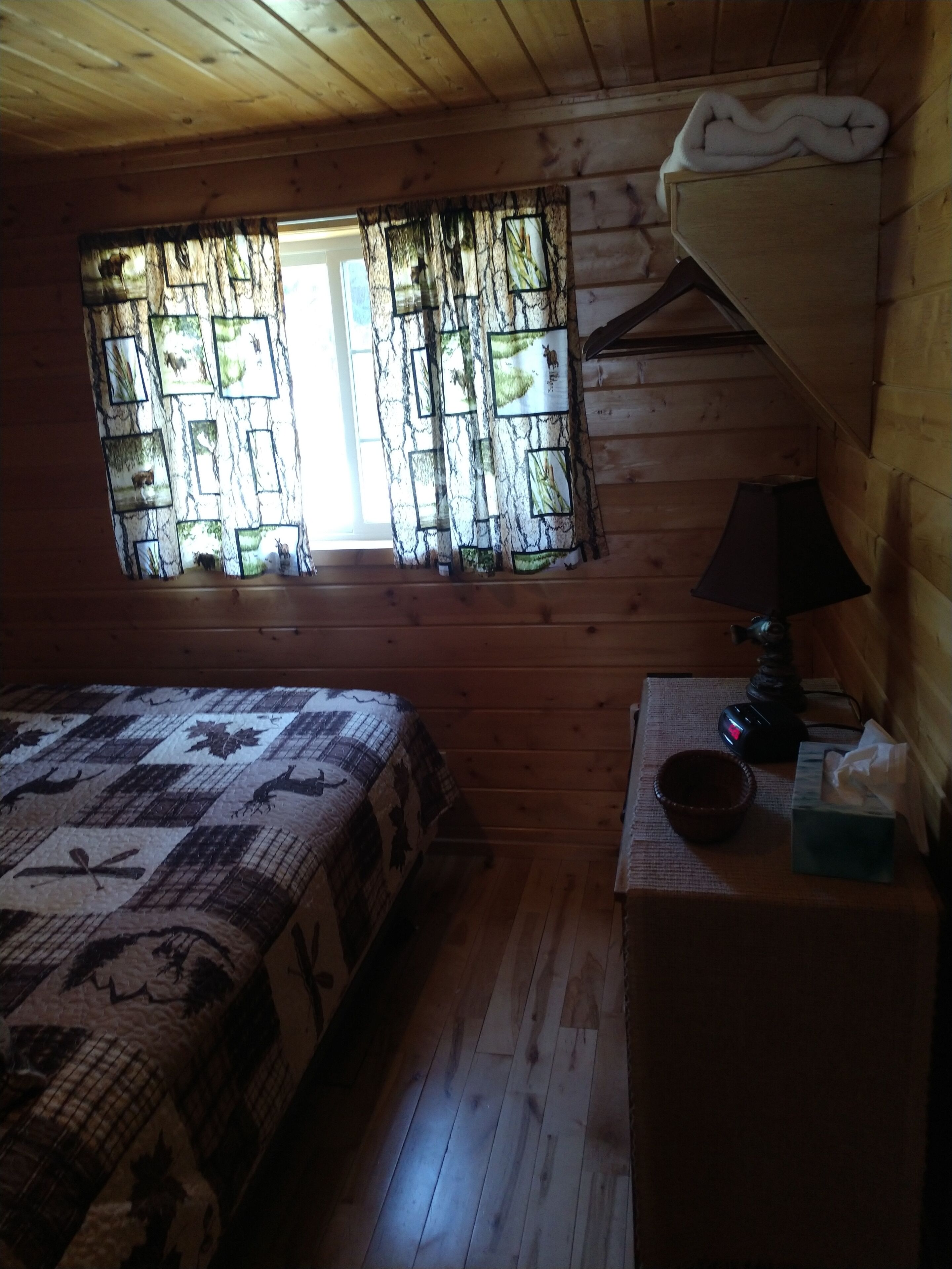 Une pièce de cabane en bois comprenant un lit, une fenêtre avec des rideaux, une lampe et une petite table avec un bol et des mouchoirs.