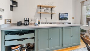 Eine Küche mit weißer Arbeitsplatte, eingebautem Spülbecken und einem Holzregal darüber. Unter der Arbeitsplatte befinden sich Schränke und zur Rechten ein Fenster.