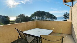 A balcony with a table and chairs, overlooking a coastal area with trees and a view of the sea.