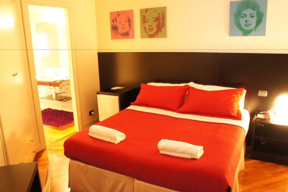 A hotel room with a large bed, red bedding, two pillows, a nightstand, and a wardrobe.