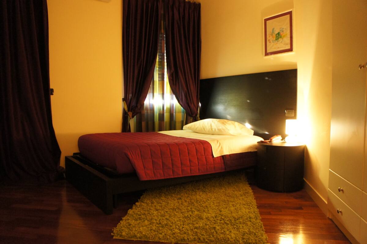 A bedroom with a bed, a nightstand, a lamp, and a window with curtains.