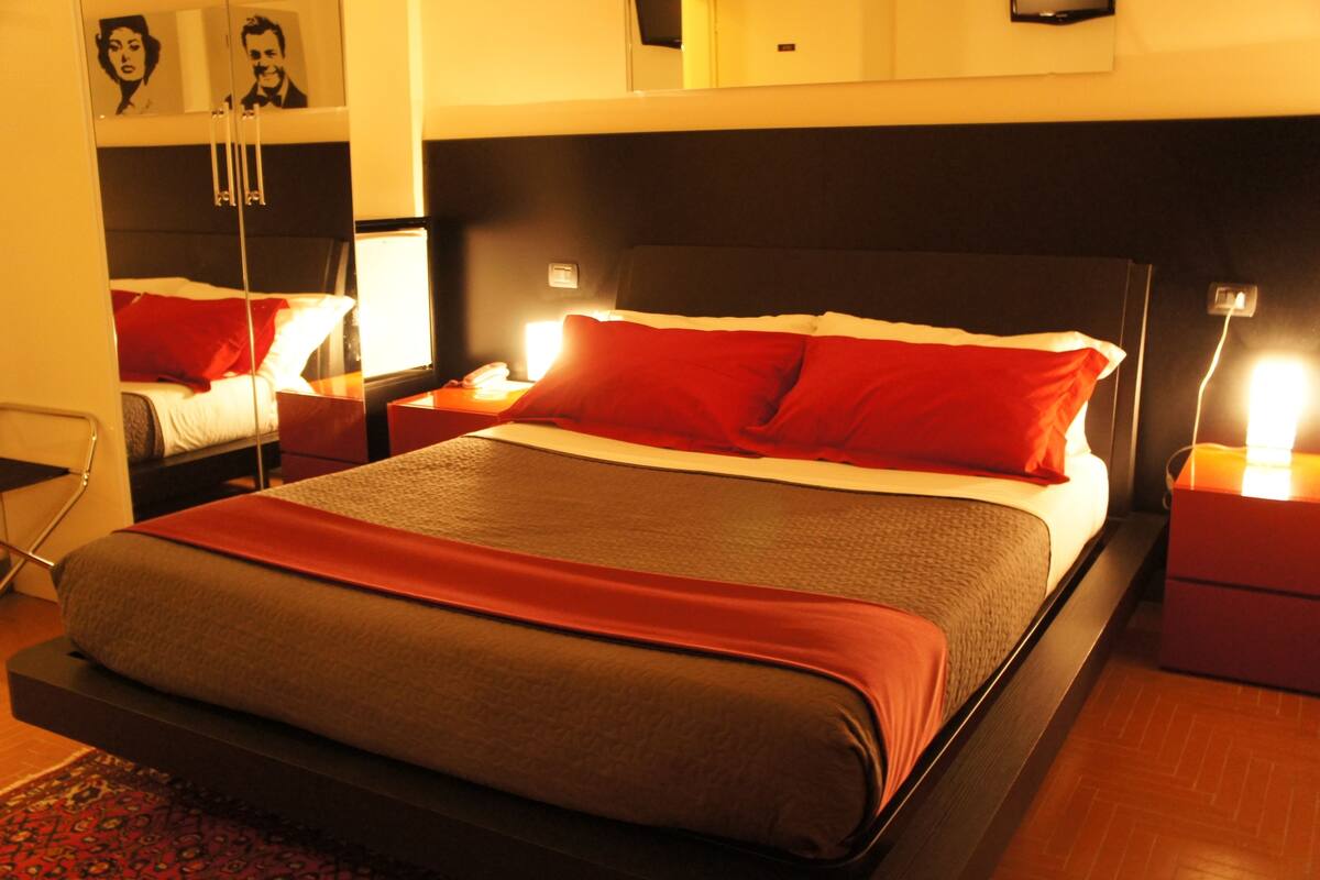 A modern hotel room with a large bed, red and grey bedding, bedside tables with lamps, and a mirror above the bed.