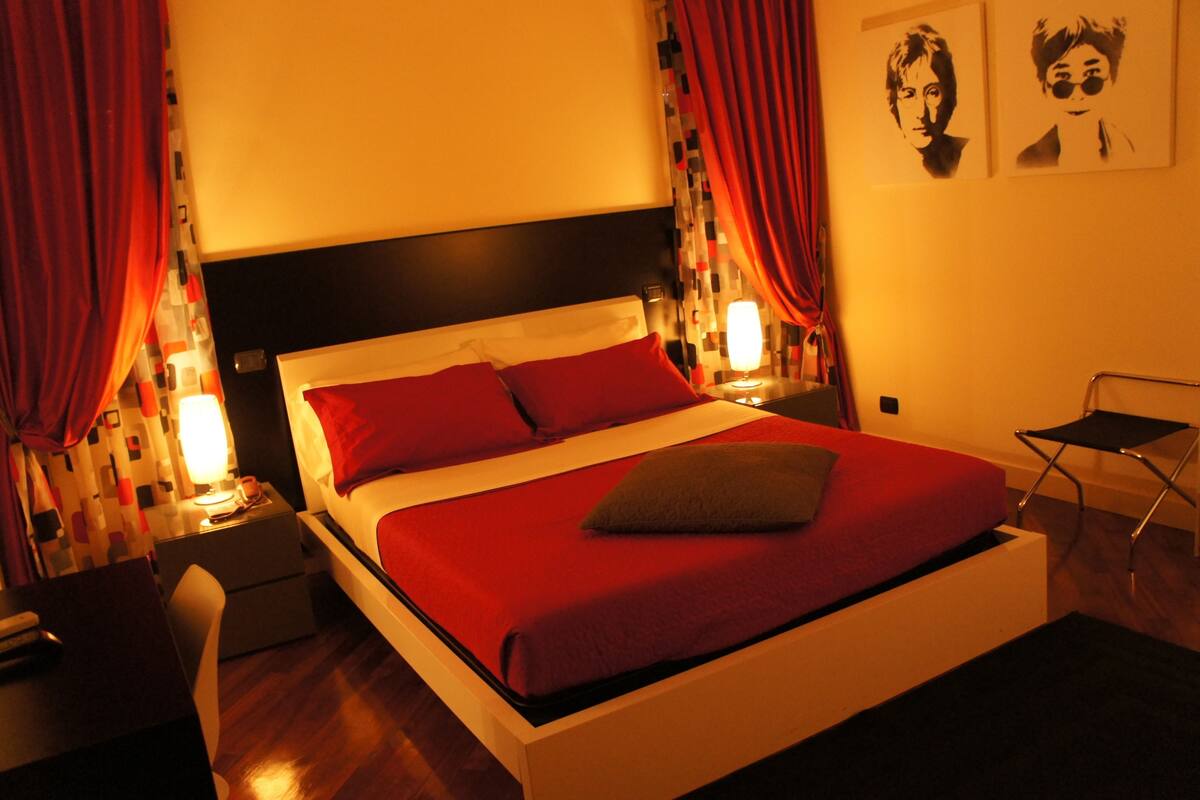A neatly made bed with red bedding, a bedside table with a lamp, and a chair.
