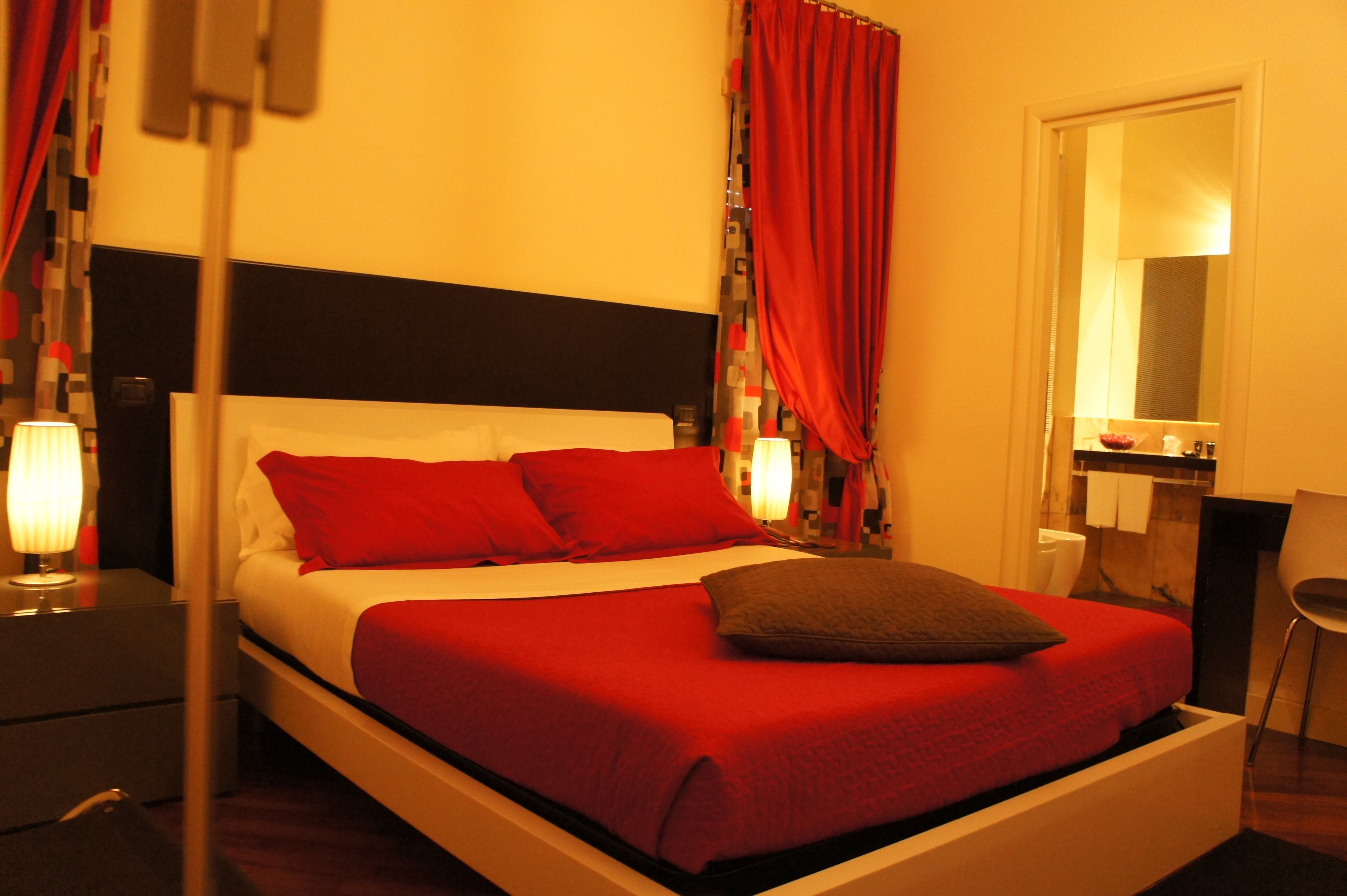 A hotel room with a bed, red curtains, a nightstand with a lamp, a mirror, and a chair.
