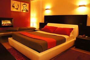 Una habitación de hotel moderna con una cama grande, ropa de cama en tonos rojo y marrón, una mesita de noche pequeña y un televisor montado en la pared.