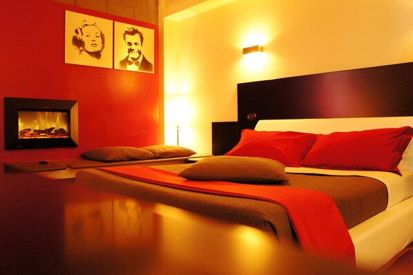Habitación de hotel con cama, almohadas rojas, cabecero, mesita de noche y chimenea empotrada.