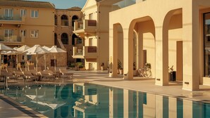 Un área de piscina con sillones de descanso y sombrillas, una piscina cristalina y edificios de varios pisos con balcones.