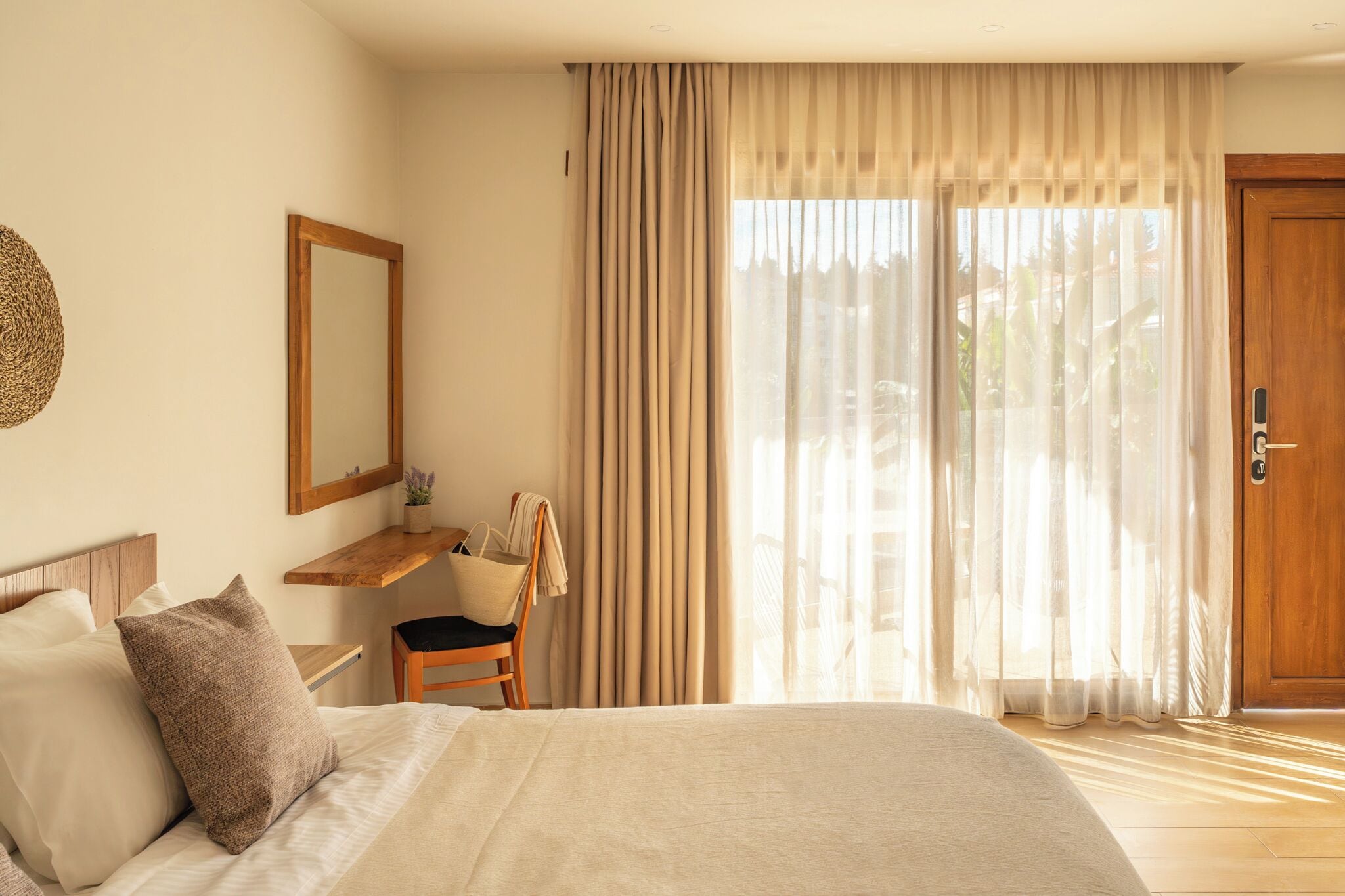 Ein Schlafzimmer mit einem Bett, einem Nachttisch aus Holz, einem Sessel, einem Schreibtisch, einem Spiegel und einer Glastür, die auf einen Balkon führt.