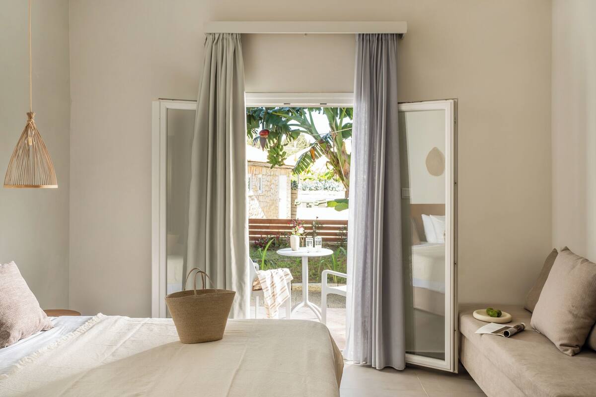 Un dormitorio con cama, una mesita de noche, una pequeña mesa con una silla afuera y una puerta corrediza de vidrio que da a un espacio exterior con plantas.