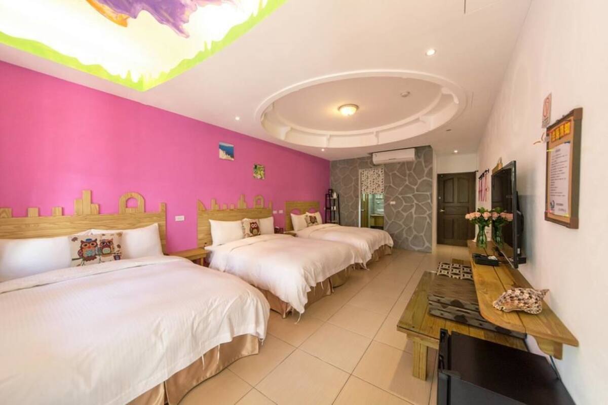 A hotel room with three beds, a wooden desk, a TV, and a stone wall.