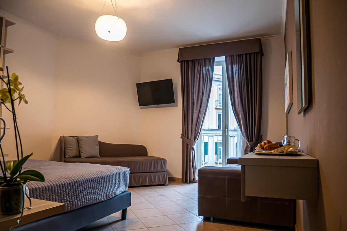 Una habitación de hotel moderna con una cama, un sofá, una mesita con una jofaina y frutas, y un televisor en la pared.