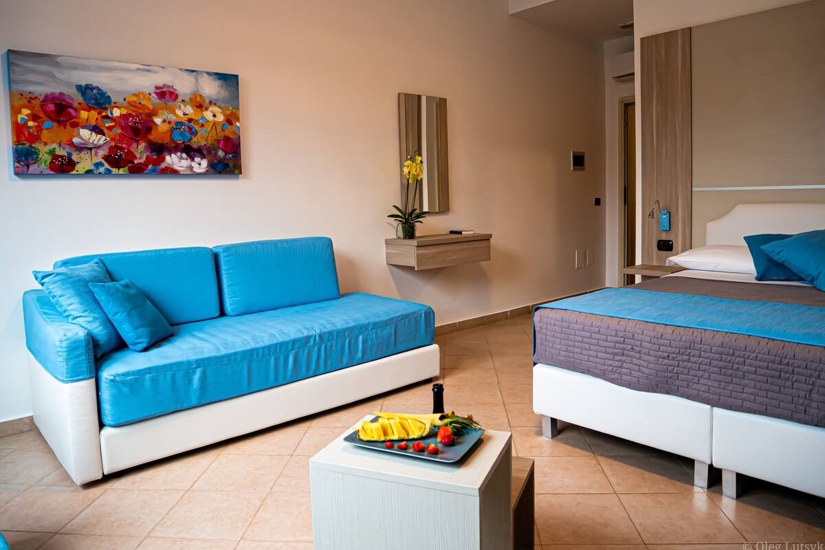 Une chambre d’hôtel moderne avec un canapé bleu, un lit recouvert d’une courtepointe bleue, une petite table avec des fruits, un tableau accroché au mur et un miroir au-dessus du canapé.
