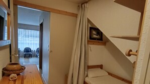 Um quarto pequeno e acolhedor com uma beliche de madeira, uma mesa pequena com uma tigela e um objeto decorativo, e uma porta que dá para outro quarto.
