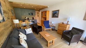 Uma sala de estar aconchegante com sofá, cadeiras, mesa de centro e cozinha ao fundo.