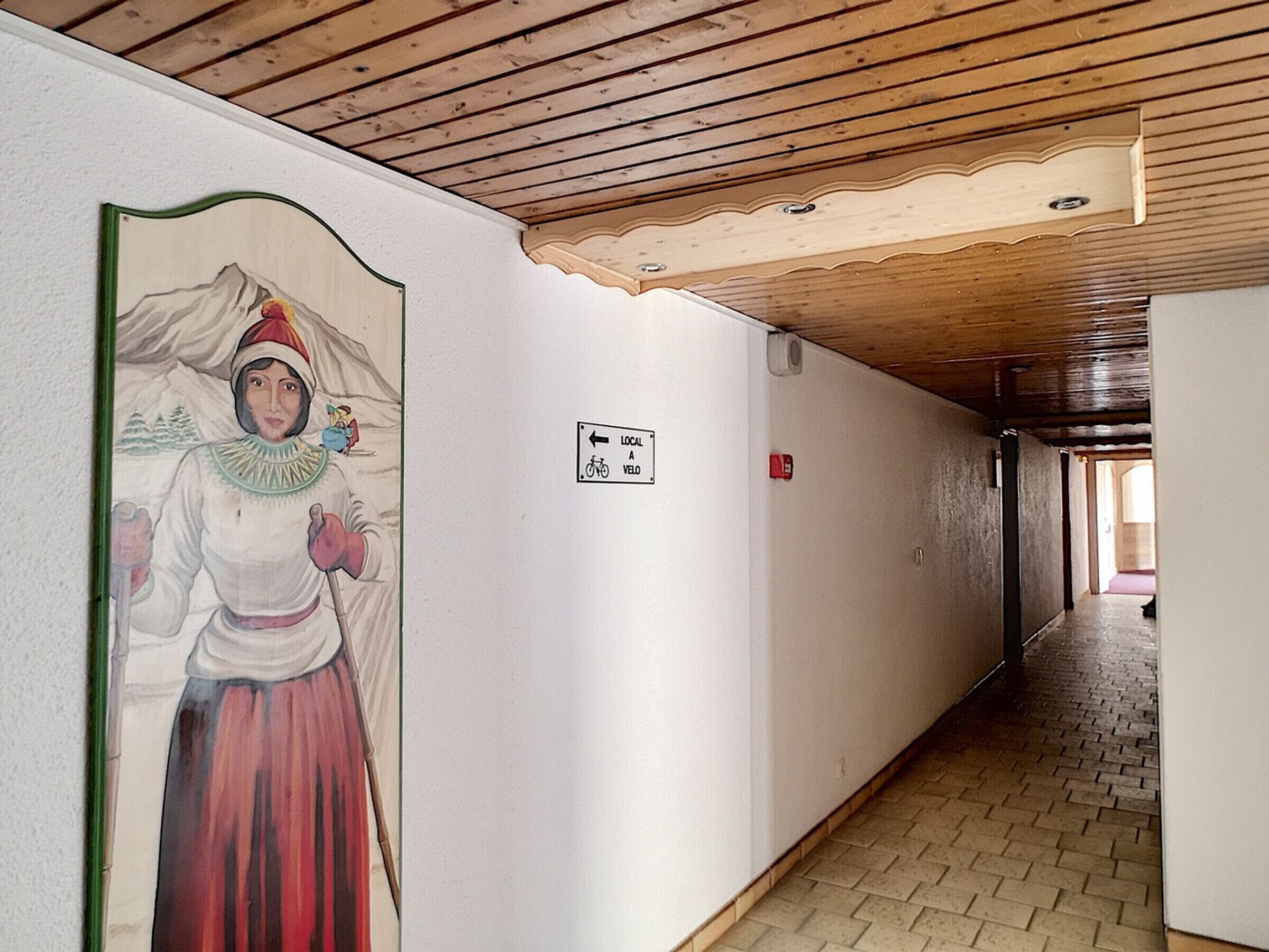 A corridor with a wooden ceiling, a mural on the left wall, and a directional sign on the right.