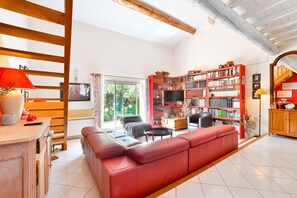 A spacious living room with a red sofa, a wooden bookshelf, and a large window providing a view of greenery. - Homerez - Enchanting Villa with Private Pool in Eyragues (Eyragues)