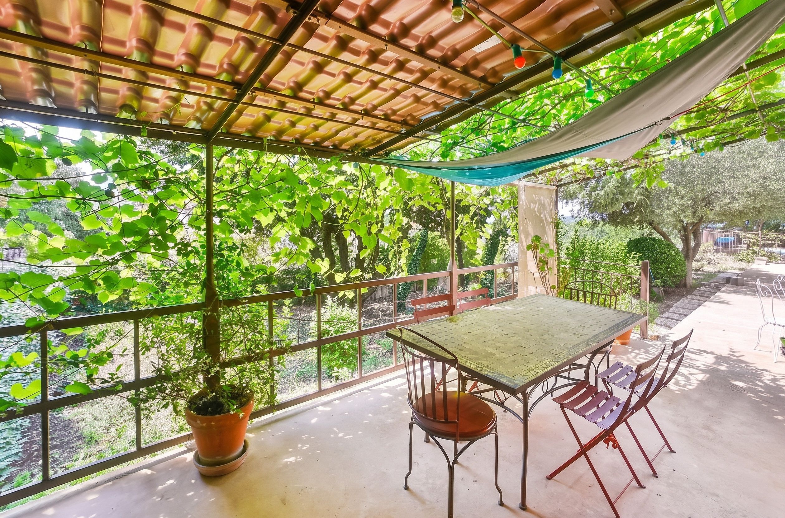 A covered outdoor seating area with a table and chairs, surrounded by greenery and a view of trees.