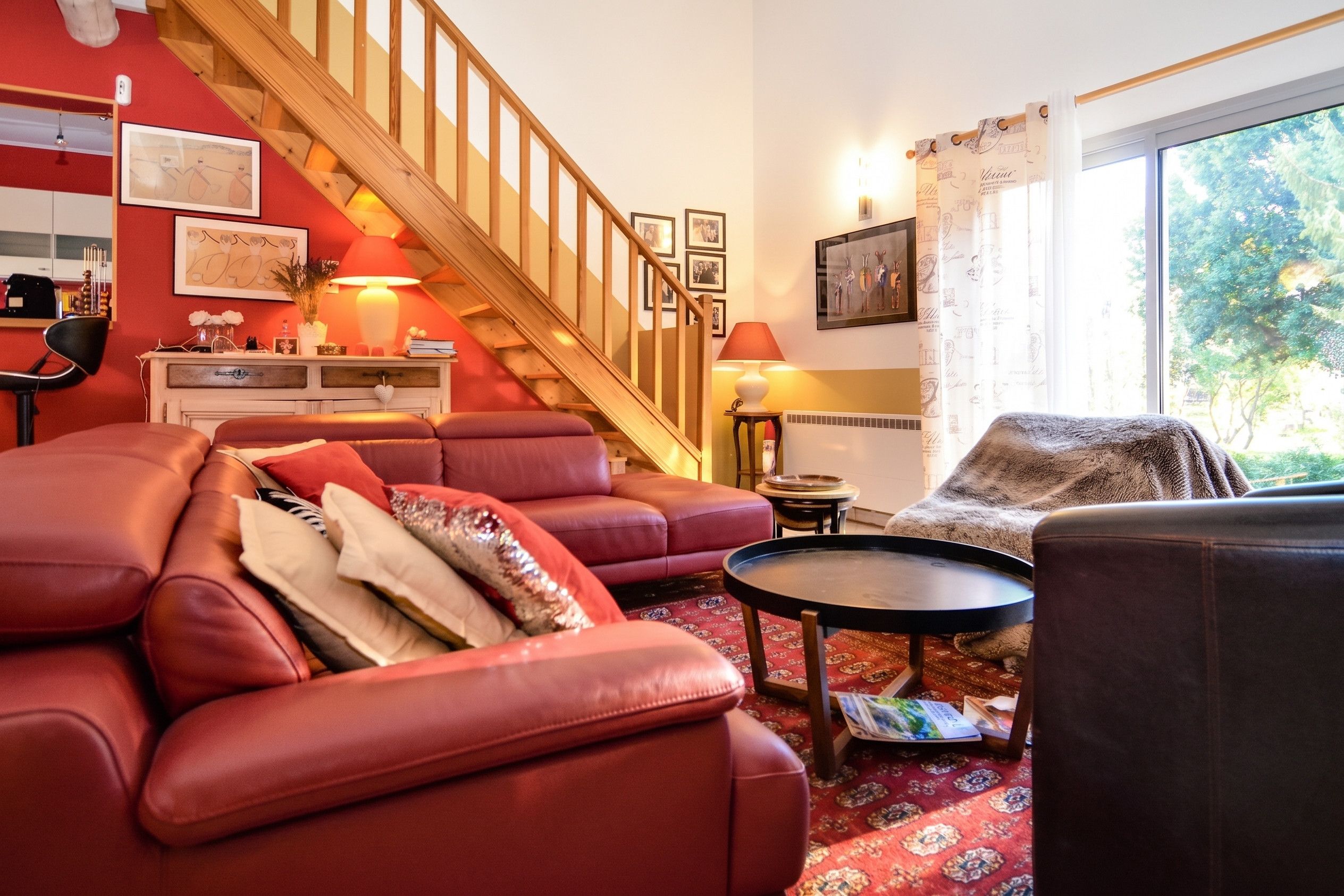 A living room with a red sofa, a wooden staircase, and a large window with curtains.
