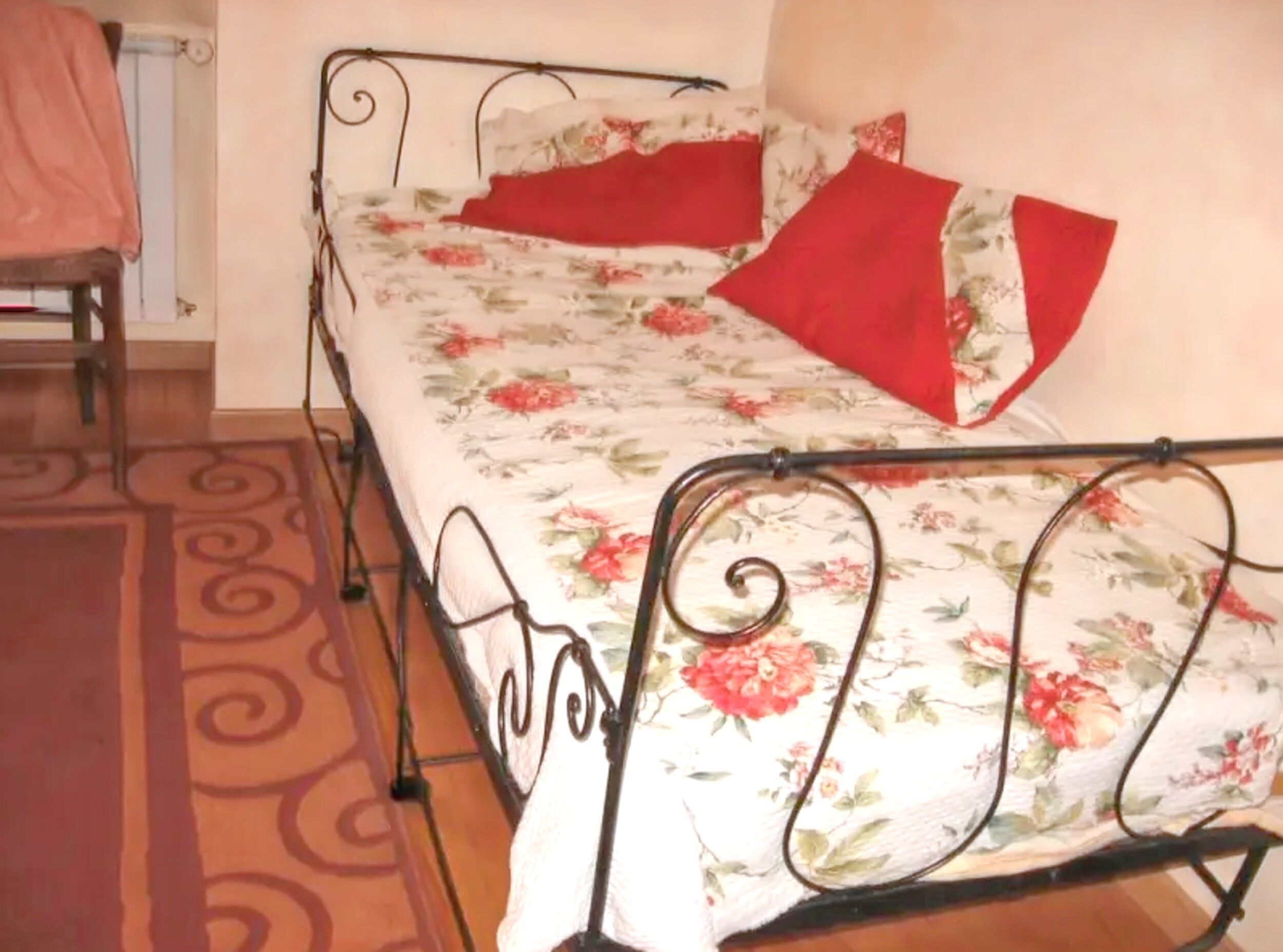A single bed with a floral bedspread and red pillows, a metal frame, and a patterned rug on a wooden floor.
