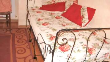 A single bed with a floral bedspread and red pillows, a metal frame, and a patterned rug on a wooden floor.
