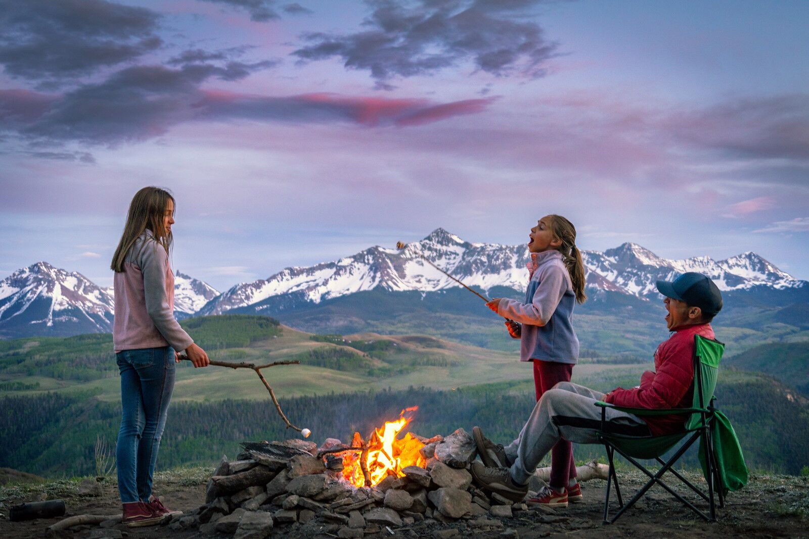 Three people around a campfire with a mountainous landscape in the background.