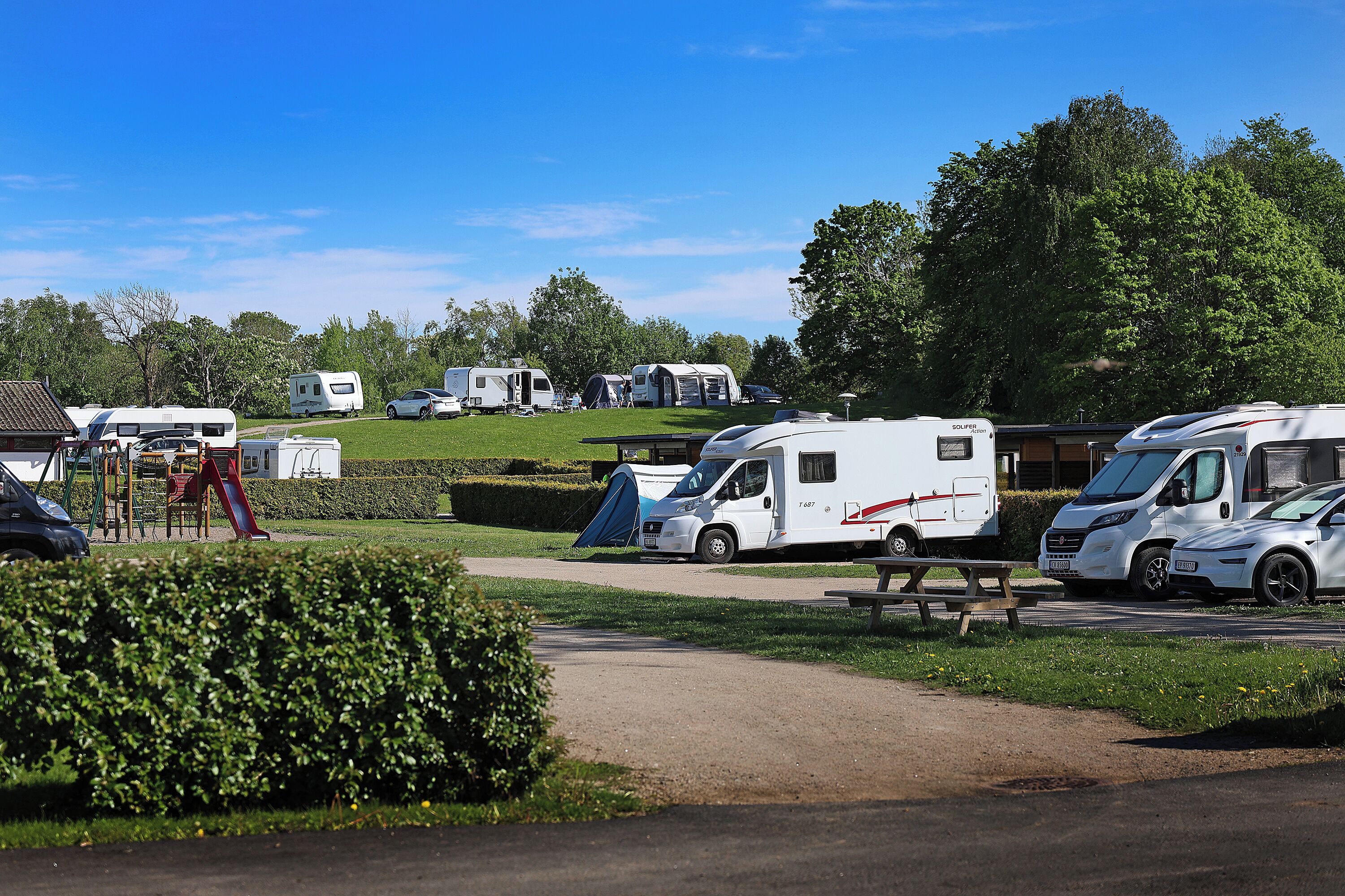 Parque para caravanas con varias caravanas estacionadas en un césped, una mesa de picnic y un parque infantil al frente.
