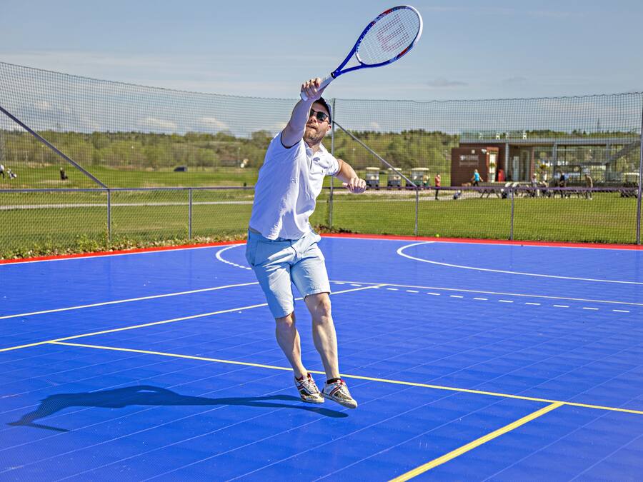 Una cancha de tenis con red, un jugador con camisa y pantaloneta blancos, y un cielo despejado al fondo.