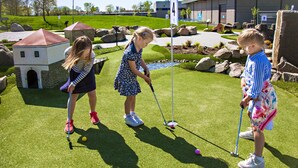 Three children playing golf on a green with a model building and rocks in the background. - Aktivitetsbyen Gamle Fredrikstad (Fredrikstad)