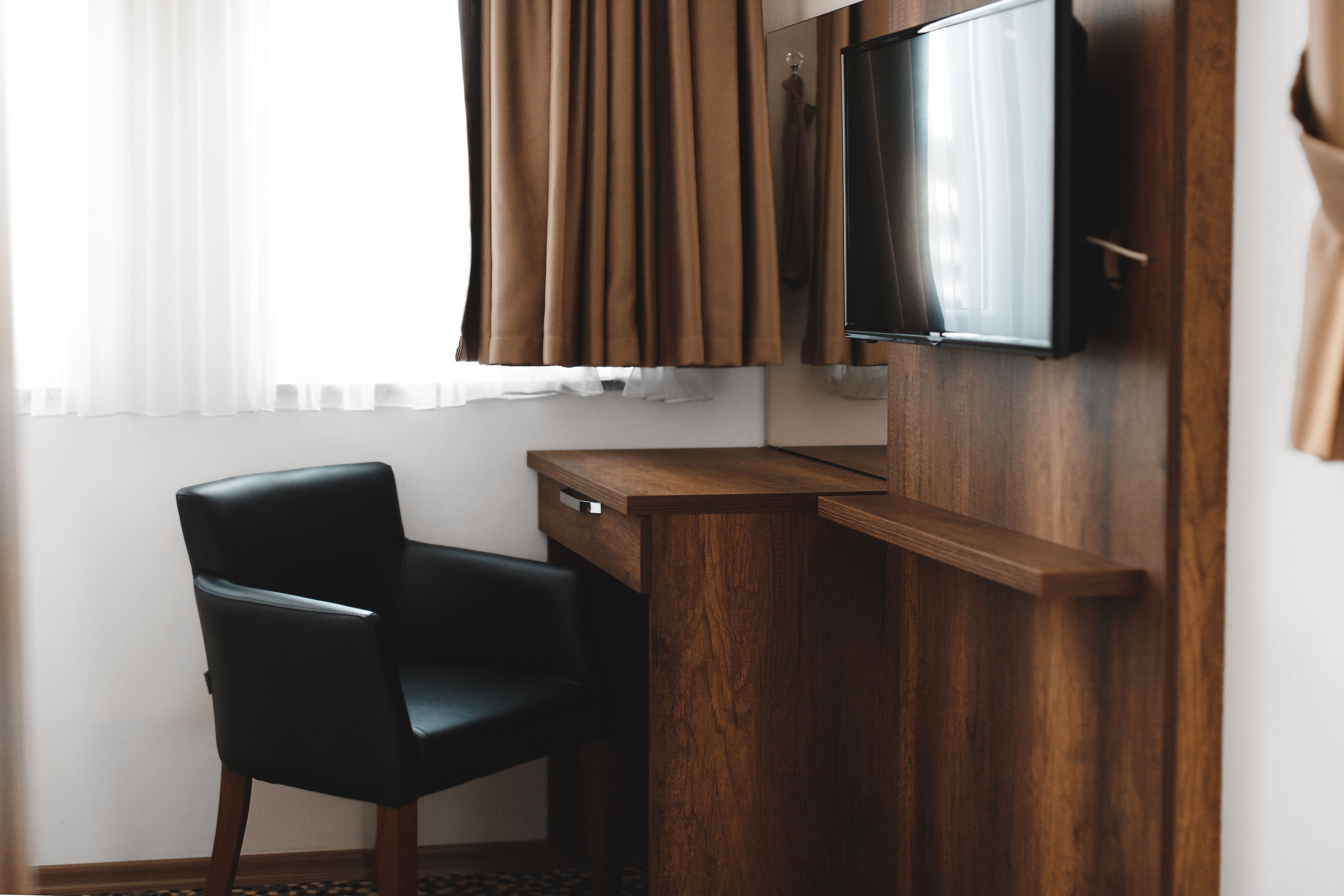 A hotel room with a wooden desk, a black leather chair, a flat-screen TV mounted on the wall, and a window with brown curtains.